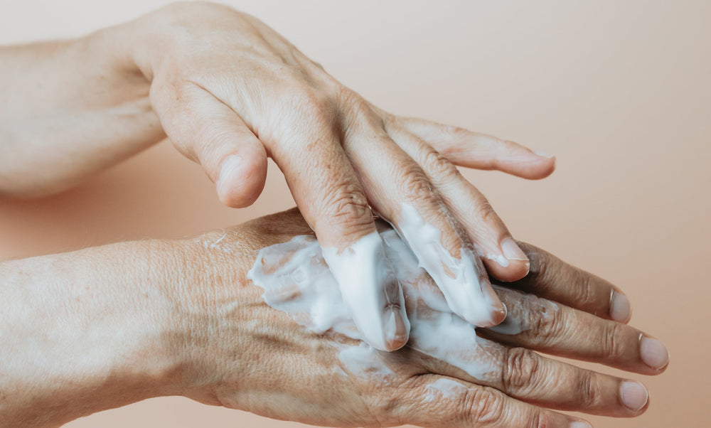 Close-up of a person's hands lathering white soap or hand wash on the back of one hand against a soft beige background.